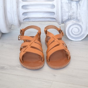 Puede incluir: Un par de sandalias de cuero color canela con m&uacute;ltiples tiras anchas en la parte superior y una correa ajustable en el tobillo con hebilla de metal. Las sandalias tienen suela plana y punta redondeada, con un acabado natural en marr&oacute;n claro.