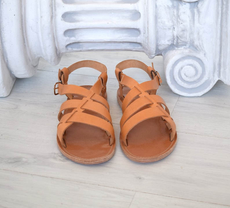 Puede incluir: Un par de sandalias de cuero color canela con m&uacute;ltiples tiras anchas en la parte superior y una correa ajustable en el tobillo con hebilla de metal. Las sandalias tienen suela plana y punta redondeada, con un acabado natural en marr&oacute;n claro.