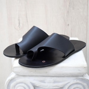 May include: A pair of black leather sandals with a toe loop design. The sandals have a minimalist style and are displayed on a white pedestal against a light wood background.