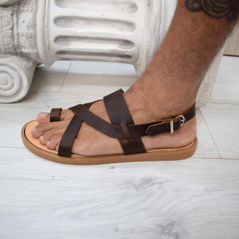 May include: A dark brown leather sandal featuring a toe loop, multiple crisscrossing straps across the top of the foot, and an adjustable ankle strap with a silver-toned metal buckle. The sandal has a flat, light tan sole with a smooth finish.