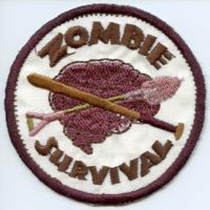 May include: A white circular patch with brown stitching around the edge. The centre of the patch features a brown brain with a crossed baseball bat and shovel. The text "Zombie Survival" is written in brown around the edge of the patch.