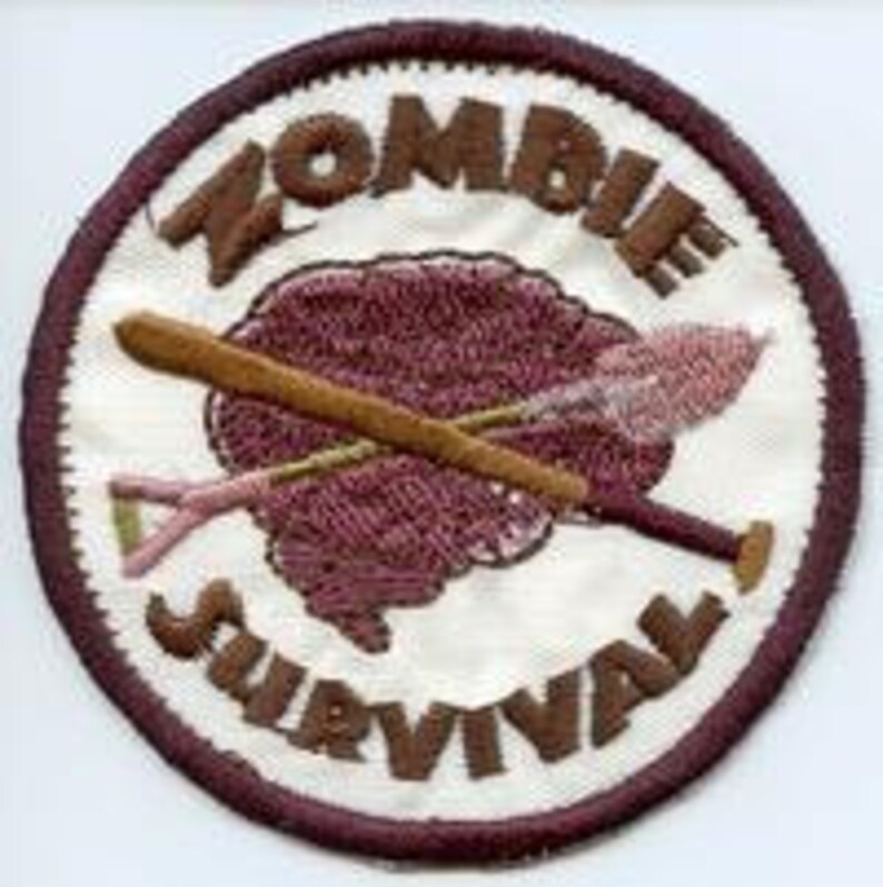 May include: A white circular patch with brown stitching around the edge. The centre of the patch features a brown brain with a crossed baseball bat and shovel. The text "Zombie Survival" is written in brown around the edge of the patch.
