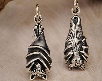 May include: Two silver bat-shaped pendants with intricate detailing. The bats are hanging upside down, with their wings and faces clearly visible. The pendants have a loop at the top for hanging and are set against a neutral background.