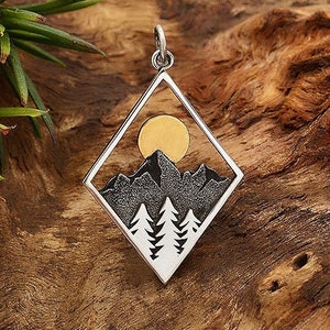 May include: A silver pendant in the shape of a diamond with a black and white mountain scene and a gold sun inside. The pendant is on a wooden surface.
