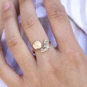 May include: A gold ring with a round, light-colored stone and a bat design. The ring is worn on a person's finger.