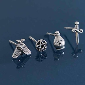 May include: Silver-toned stud earrings featuring various designs: a moth, a pentagram, a pouch with crosses, and a sword. The earrings are displayed on a dark blue surface, showcasing their intricate details and reflective quality.
