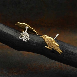 May include: A pair of gold bird stud earrings. The earrings are made of metal and feature a detailed design of a bird with its wings spread. The earrings are shown on a dark wooden surface.