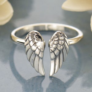 May include: A silver-colored ring featuring a detailed angel wing design. The wings are positioned to appear as if they are embracing the finger. The ring has a polished finish and is set against a soft, blurred background.