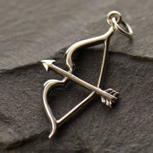 May include: A silver charm in the shape of a bow and arrow. The arrow is pointed and the bow is curved into a heart shape.