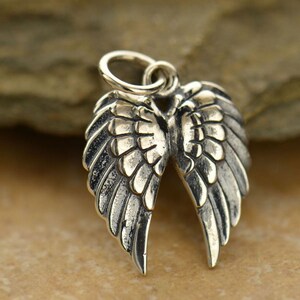 May include: A detailed silver-colored pendant in the shape of angel wings. The wings are open, with intricate feather detailing. A small loop is at the top for attaching to a chain or bracelet. The pendant is likely made of sterling silver.