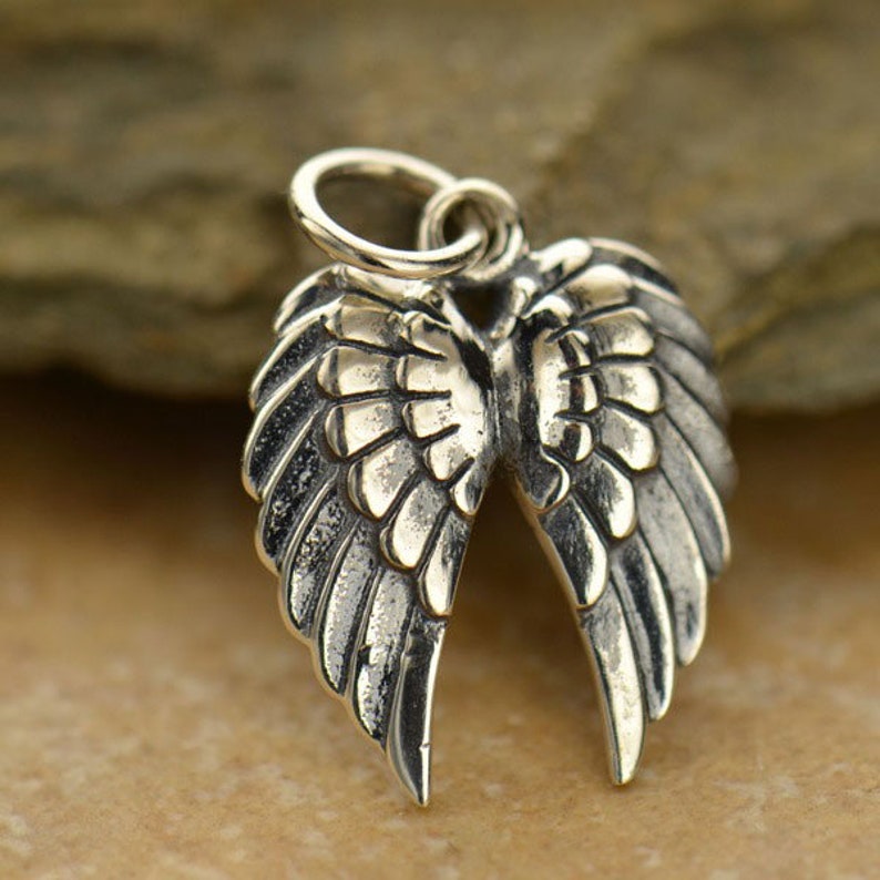 May include: A detailed silver-colored pendant in the shape of angel wings. The wings are open, with intricate feather detailing. A small loop is at the top for attaching to a chain or bracelet. The pendant is likely made of sterling silver.