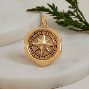 May include: A gold-colored pendant featuring a compass rose design. The pendant has a circular shape with a raised star in the center, surrounded by a detailed border. The pendant is suspended from a small loop at the top.