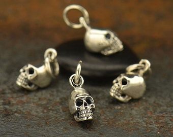 May include: Four silver skull charms with a loop for attaching to a necklace or bracelet. The charms are detailed with eye sockets and a nose.