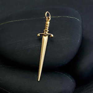 May include: A gold-colored sword charm pendant with a detailed design. The sword has a pointed tip and a cross guard.