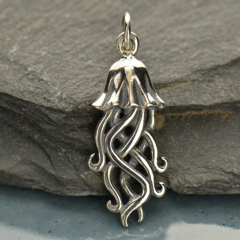 May include: A silver charm in the shape of a jellyfish with a detailed, intricate design. The charm has a bell-shaped top and long, flowing tentacles.