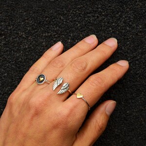 May include: A hand wearing four silver rings. One ring features a gold heart, another has silver wings, a third has a gold heart, and the last has a gold heart inside a black circle.