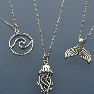 May include: Three silver necklaces with different ocean-themed pendants: a wave in a circle, a jellyfish, and a mermaid tail.