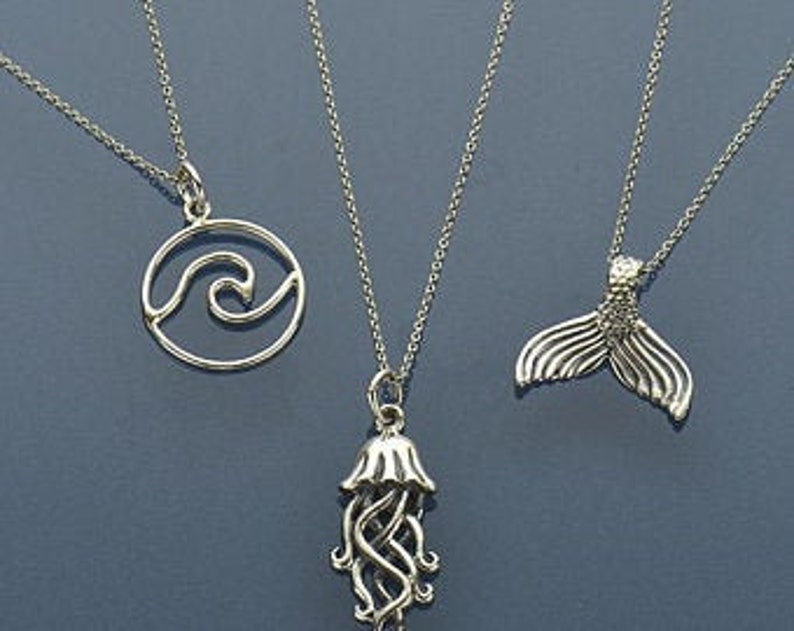 May include: Three silver necklaces with different ocean-themed pendants: a wave in a circle, a jellyfish, and a mermaid tail.