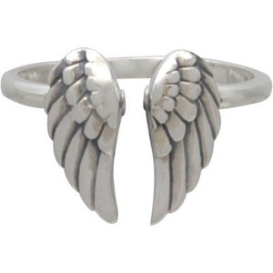 May include: A silver-colored ring featuring a design of two open wings. The wings are detailed with layered feather patterns, creating a textured appearance. The ring's band is a simple, smooth design.