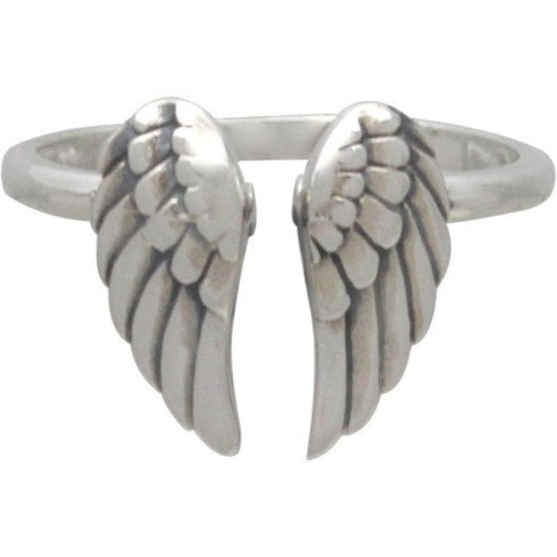 May include: A silver-colored ring featuring a design of two open wings. The wings are detailed with layered feather patterns, creating a textured appearance. The ring's band is a simple, smooth design.