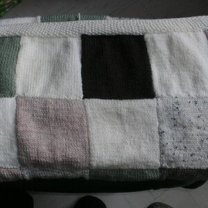 May include: A handmade, patchwork blanket featuring a variety of neutral colors. The blanket is composed of squares in shades of white, green, brown, and pink. The edges are finished with a white knitted border. The blanket is a cozy, handcrafted item.