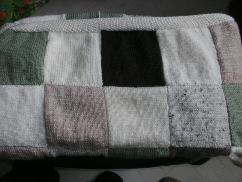 May include: A handmade, patchwork blanket featuring a variety of neutral colors. The blanket is composed of squares in shades of white, green, brown, and pink. The edges are finished with a white knitted border. The blanket is a cozy, handcrafted item.