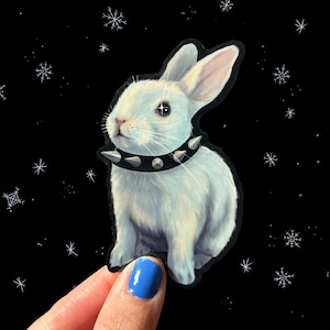 Könnte beinhalten: Ein weißes Kaninchen mit einem schwarzen, mit Nieten besetzten Halsband vor einem schwarzen Hintergrund mit weißen Schneeflocken.