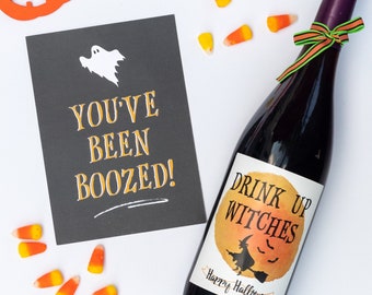 May include: A black card with the text "You've Been Boozed!" and a white ghost illustration. A bottle of wine with a label that says "Drink Up Witches" and a witch riding a broomstick. The bottle is surrounded by orange and white candy corn.