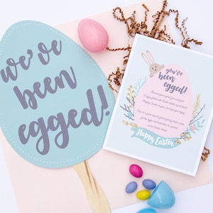 May include: A blue and white egg-shaped card with the text "we've been egged!" and a smaller card with the text "you've been egged!" with a bunny peeking out from behind the text. The smaller card has a floral design and the text "Happy Easter".