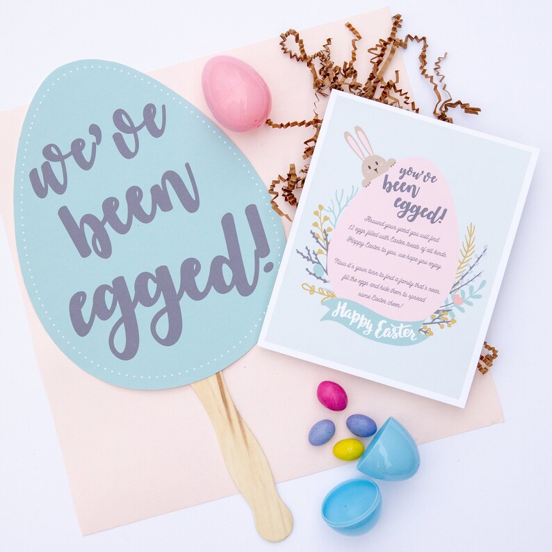 May include: A blue and white egg-shaped card with the text "we've been egged!" and a smaller card with the text "you've been egged!" with a bunny peeking out from behind the text. The smaller card has a floral design and the text "Happy Easter".