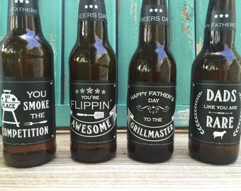 May include: Four brown glass beer bottles with black and white labels. The labels have different sayings on them, including "You're Flipping Awesome", "You Smoke the Competition", "Happy Father's Day to the Grillmaster", and "Dads Like You Are Rare".