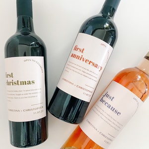 Pode incluir: Tr&ecirc;s garrafas de vinho com r&oacute;tulos personalizados. Os r&oacute;tulos s&atilde;o brancos com texto preto e apresentam as palavras "primeiro natal", "primeiro anivers&aacute;rio" e "s&oacute; porque". Cada r&oacute;tulo tamb&eacute;m inclui os nomes "Christina + Christopher" e a data "11.05.22".
