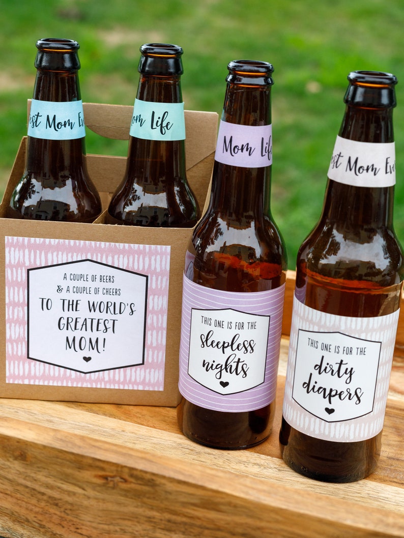 May include: Four brown glass beer bottles with labels. The labels have a pink and white pattern and black text. The text on the labels reads "To the world's greatest mom!", "This one is for the sleepless nights", "This one is for the dirty diapers", and "Mom Life".