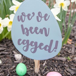 May include: A blue egg-shaped sign with the text "we've been egged!" in purple lettering. The sign is stuck in the ground with a wooden stick. There are two plastic Easter eggs on the ground, one green and one pink.