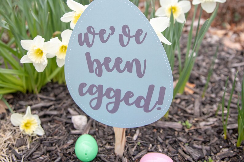 May include: A blue egg-shaped sign with the text "we've been egged!" in purple lettering. The sign is stuck in the ground with a wooden stick. There are two plastic Easter eggs on the ground, one green and one pink.
