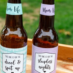 May include: Two brown glass bottles with labels that say "Mom Life". The bottle on the left has a light blue label with white brushstrokes and the text "This one is for the drool & spit up". The bottle on the right has a purple label with white stripes and the text "This one is for the sleepless nights".