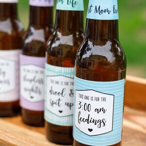 May include: Four brown glass bottles with blue, green, purple, and white labels. The labels have text that reads "This one is for the drool & spit up", "This one is for the 3:00 am feedings", "First Mom Ever", and "Mom Life".