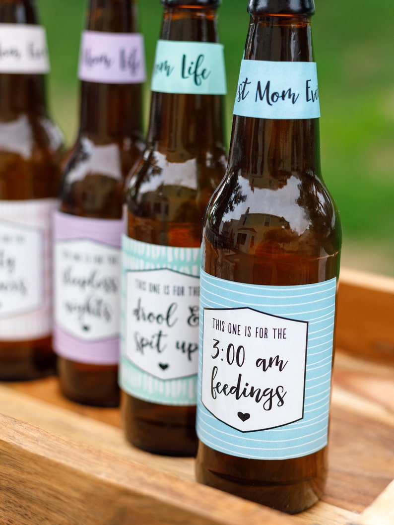 May include: Four brown glass bottles with blue, green, purple, and white labels. The labels have text that reads "This one is for the drool & spit up", "This one is for the 3:00 am feedings", "First Mom Ever", and "Mom Life".