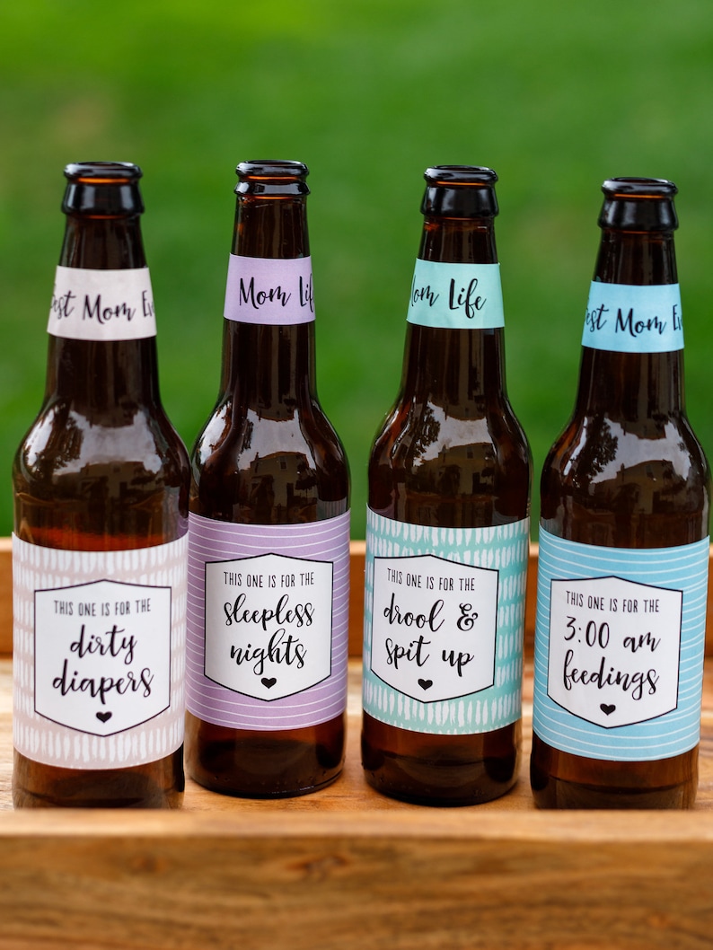 May include: Four brown glass beer bottles with white labels. Each label has a different phrase related to motherhood, such as "dirty diapers", "sleepless nights", "drool & spit up", and "3:00 am feedings".
