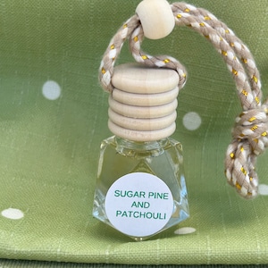 以下が含まれることがあります： 白いラベルに「シュガーパインとパチュリ」と書かれた小さなガラス瓶。瓶はベージュとゴールドの紐に木製のビーズが付いた状態で吊り下げられている。