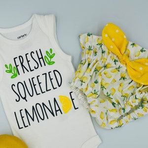 Könnte beinhalten: Ein weißer Baby-Body mit dem Text "FRESH SQUEEZED LEMONADE" und einer Zitronengrafik, kombiniert mit einer Windel mit Zitronenmuster und einem gelben Schleifen-Stirnband. Eine ganze Zitrone ist ebenfalls vorhanden.
