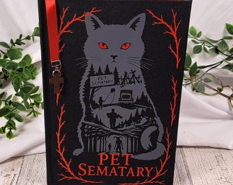 May include: A black hardcover book titled Pet Sematary featuring a gray cat illustration with red eyes, surrounded by a red branch border. A red ribbon bookmark with a small cross charm hangs from the top edge of the book cover.