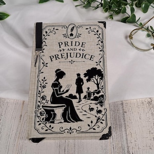May include: A rectangular book titled Pride and Prejudice with a beige fabric cover. The front features a black silhouette illustration of a person sitting on a bench and another standing in a garden, framed by decorative floral borders and corner accents.