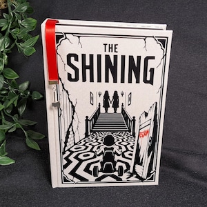 May include: A hardcover book titled The Shining with a white cover featuring black graphic illustrations of a child on a tricycle, a patterned carpet, and two figures on a staircase. A red ribbon bookmark with a small silver key charm is attached.