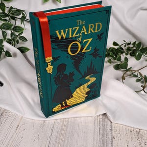 May include: A hardcover book titled The Wizard of Oz with a teal cover featuring yellow text, a yellow brick road, and a silhouette of a person holding a dog. A red ribbon bookmark with a small gold-colored charm hangs from the top of the book.