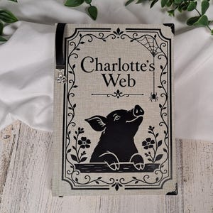 May include: A rectangular book with a light beige linen-textured cover featuring a black graphic of a pig and the text Charlotte's Web. The design includes a black decorative border, a spider web, a small spider, and a black ribbon bookmark with a charm.