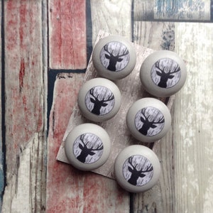 May include: Six grey cabinet knobs featuring a black deer head design on a white and grey background. The knobs are arranged on a light-coloured wooden surface, ideal for adding a rustic touch to furniture.