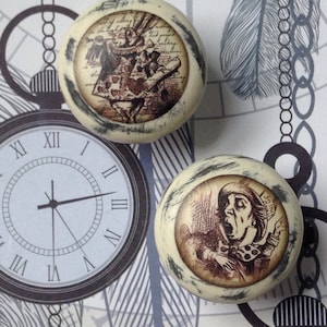 May include: Two cream-coloured cabinet knobs featuring Alice in Wonderland illustrations. One knob depicts the White Rabbit, the other the Mad Hatter. The knobs have a distressed, vintage appearance with dark grey edging. The background includes a clock and chain design.