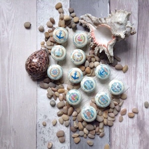 May include: A selection of nautical-themed spheres featuring anchors, sailboats, and lighthouses. The spheres are displayed on a bed of pebbles, with a seashell and a patterned shell in the background. The colour scheme includes blue, white, and beige, evoking a seaside theme.