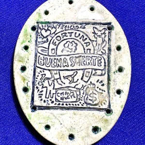 May include: A white oval ceramic charm with holes around the edge. The charm has a green glaze and a black design with the words "Buena Suerte" and "Fortuna" in the center.